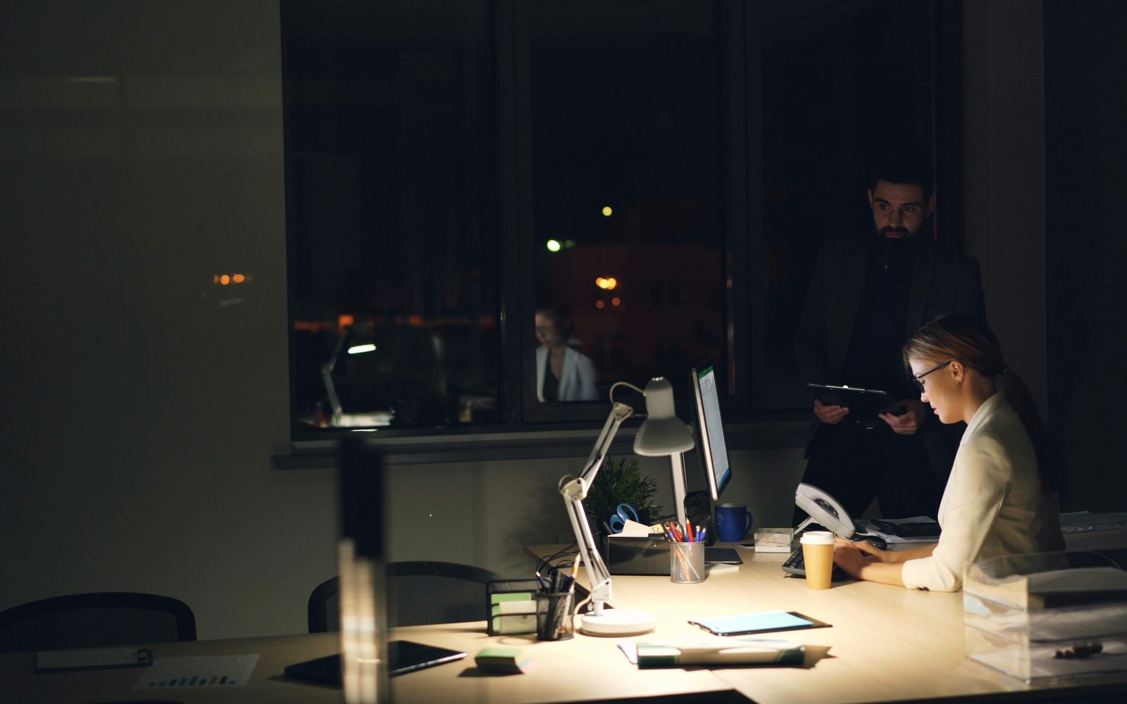 An office at night with one lit desk, a seated engineer and a colleague standing with a tablet